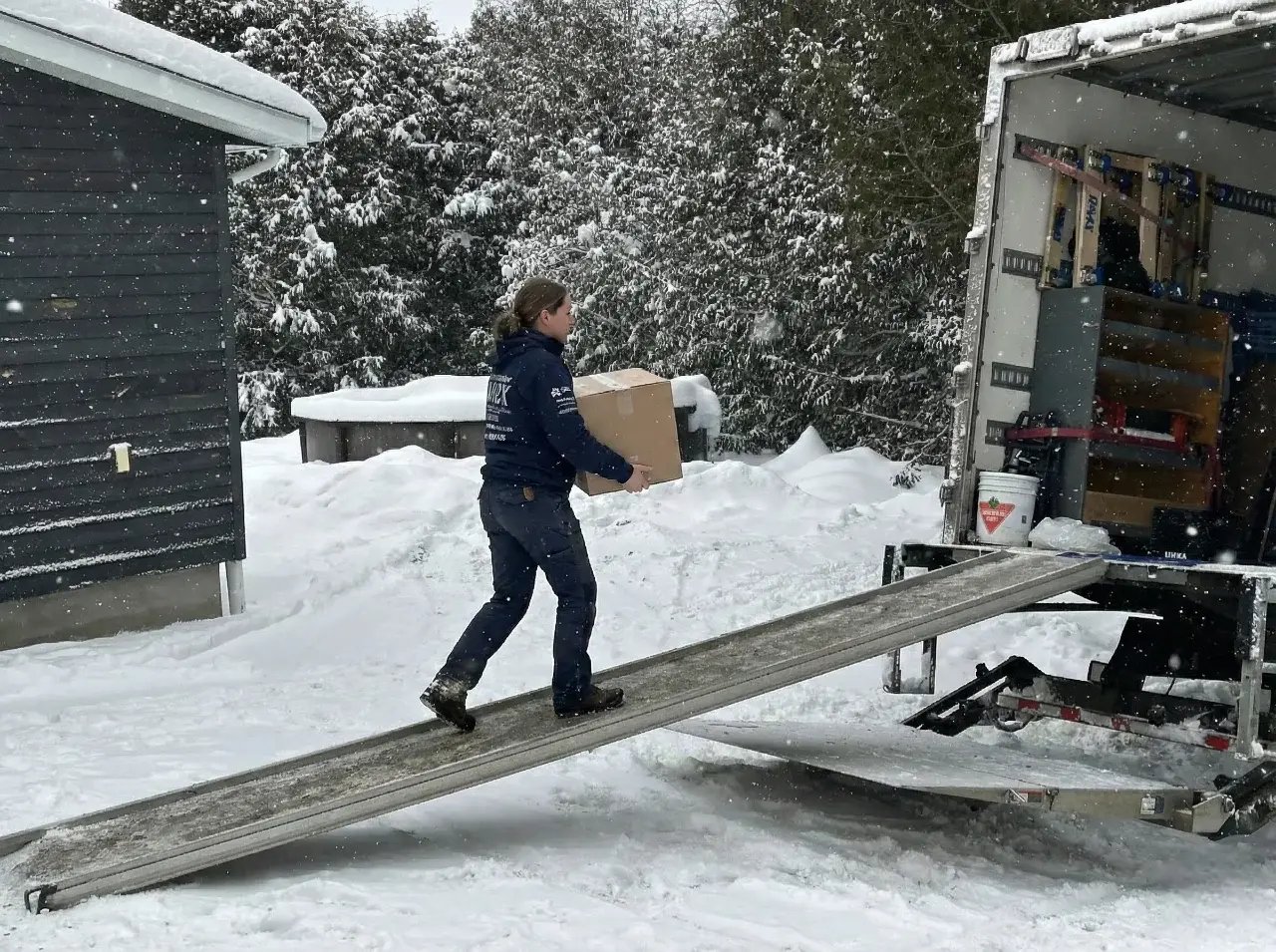 Une déménageuse porte un carton et monte la rampe d'un camion de déménagement sous la neige. Une déménageuse porte un carton et monte la rampe d'un camion de déménagement sous la neige.