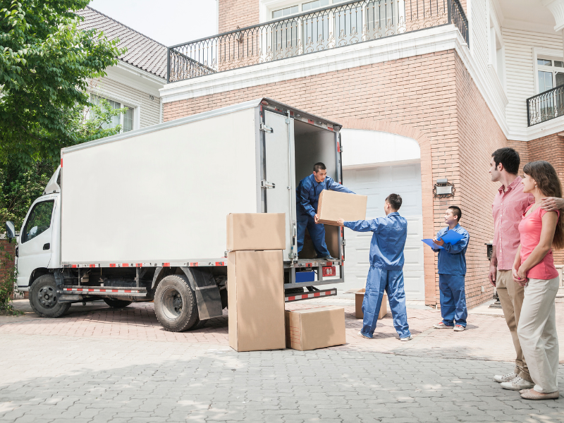 Des déménageurs déchargeant des cartons d'un camion devant une maison en présence d'un couple satisfait.