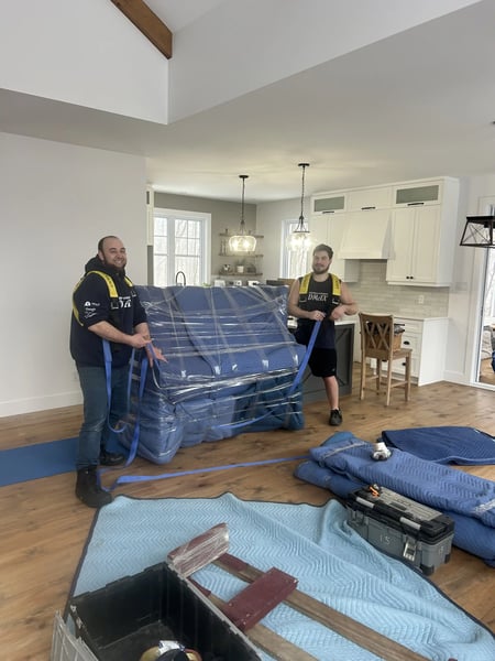 Best Mover Montreal: Two DMAX movers transporting a wrapped and protected piece of furniture in a living room, using safety equipment for a professional move.