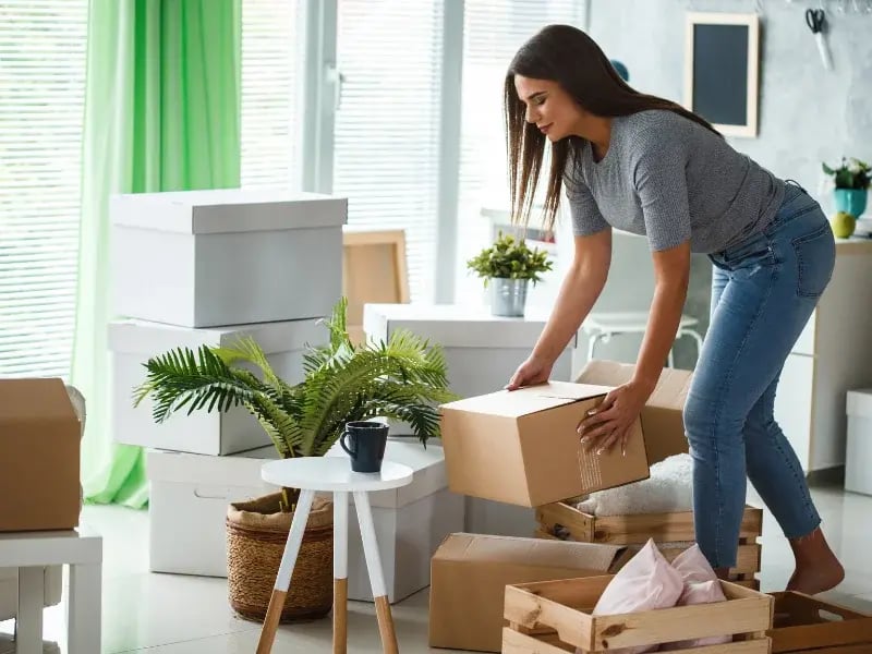 Femme organisant des boîtes en carton dans un salon bien éclairé avec des plantes et des meubles minimalistes.