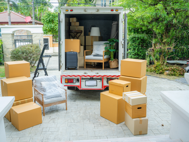 Chargement de meubles et de boîtes dans un camion de déménagement, ouvert et stationné devant une maison, avec divers articles ménagers et boîtes empilées à l'extérieur.