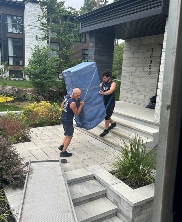 Deux déménageurs transportant un meuble enveloppé dans une couverture bleue sur les marches d'une maison moderne.