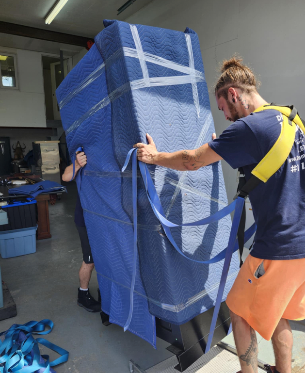 Deux déménageurs transportant un meuble enveloppé dans une couverture de protection bleue à l'aide de sangles.