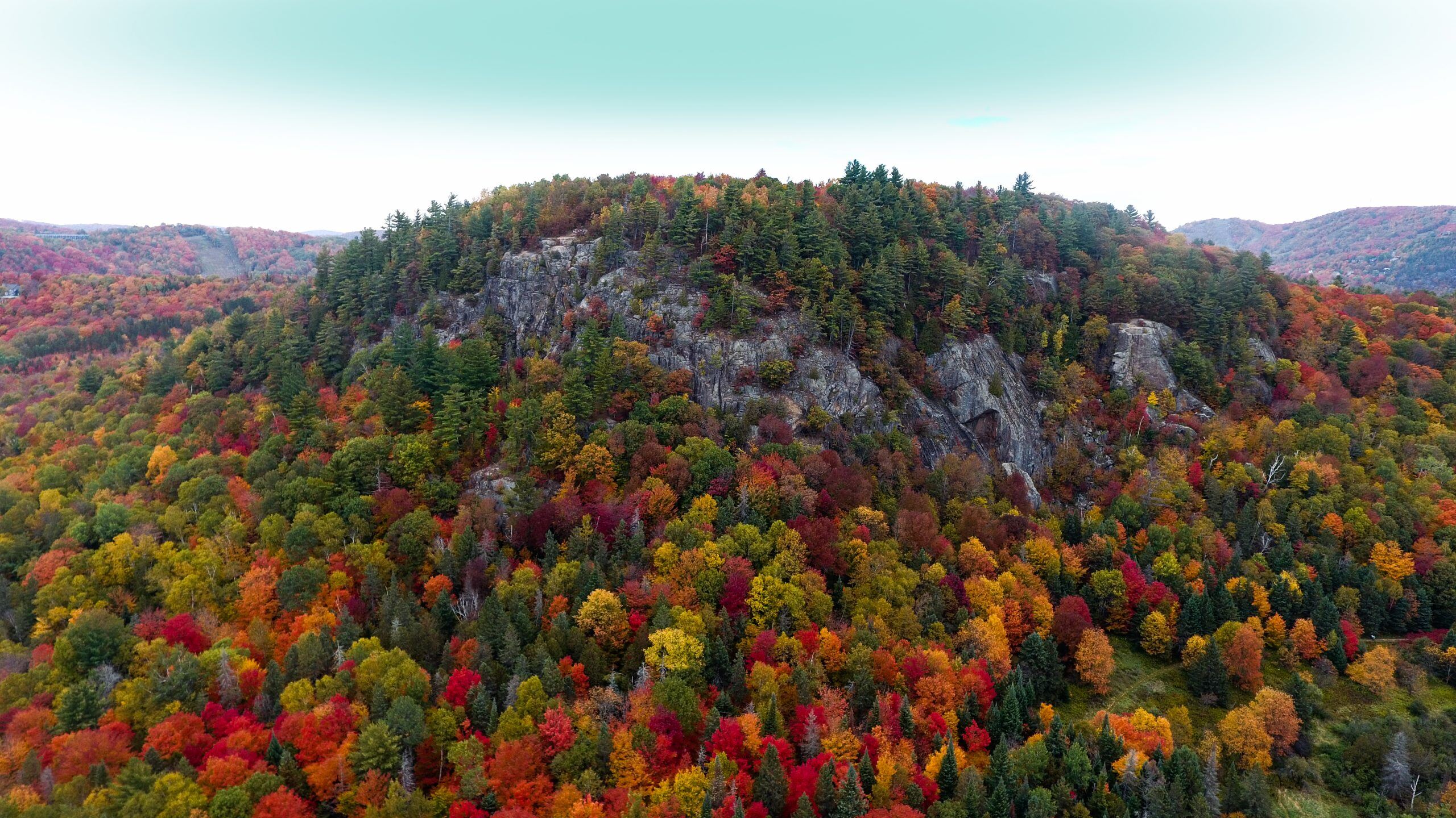 Vue aérienne du feuillage automnal couvrant les collines et falaises de Saint-Sauveur, Québec, présentant un mélange d'arbres rouges, orange, jaunes et verts sous un ciel dégagé.