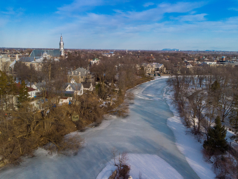 Moving to Terrebonne: Aerial view of Terrebonne in Quebec during winter, showing a frozen river, houses surrounded by bare trees, and a church with a steeple visible on the horizon.