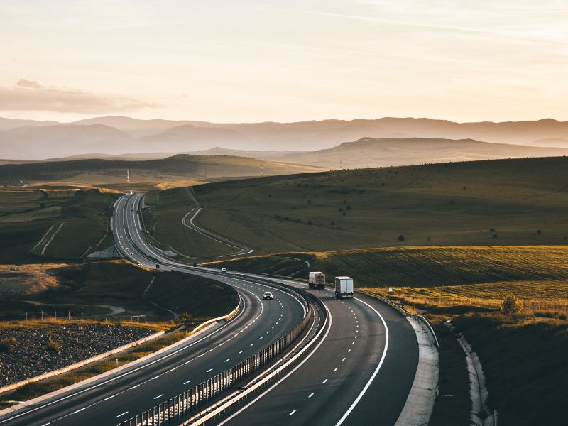 Winding highway with trucks and cars, surrounded by green fields and mountains at sunset.