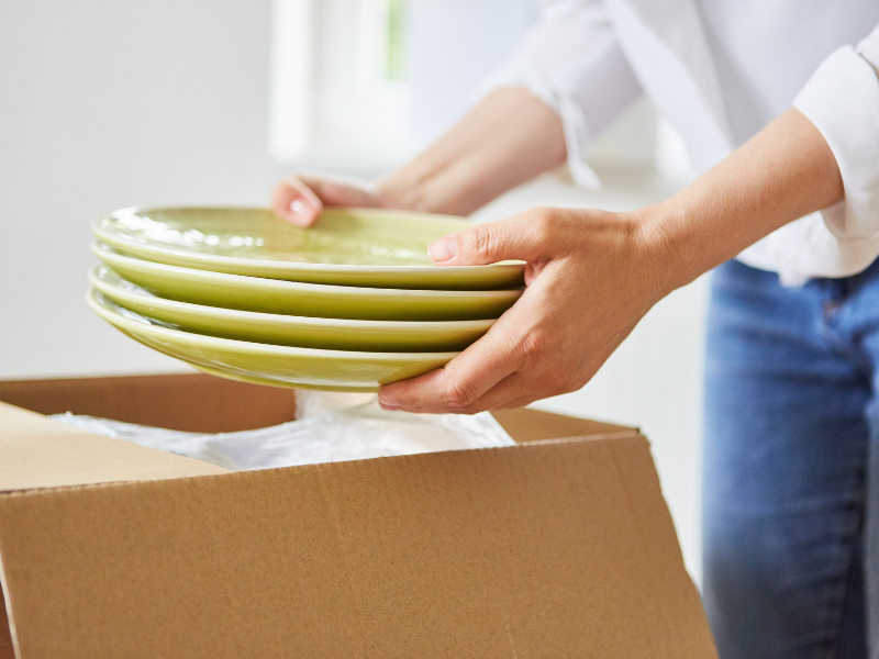 Person carefully packing green plates into a moving box, ensuring their protection with bubble wrap.