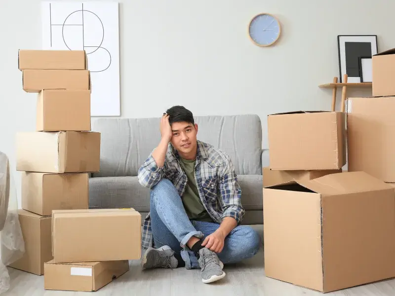 Man sitting on the floor, surrounded by moving boxes in a living room, looking tired.